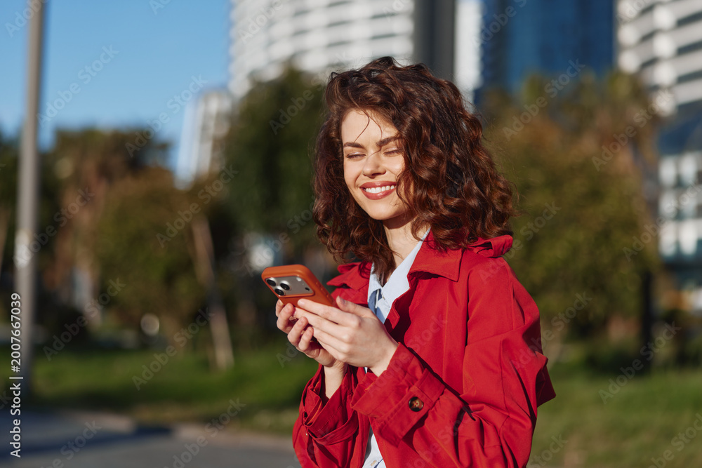 Fototapeta premium Smiling woman with curly hair wearing a red jacket checks her smartphone in a sunny park. Casual lifestyle, outdoor moment, bright mood, candid urban greenery and vitality.