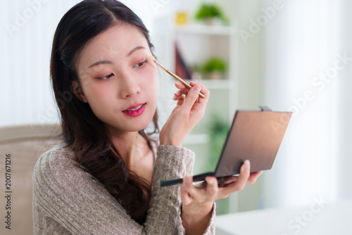 Asian women applying eyeshadow makeup with brush looking at mirror doing morning beauty routine at home using cosmetics products compact palette for skincare