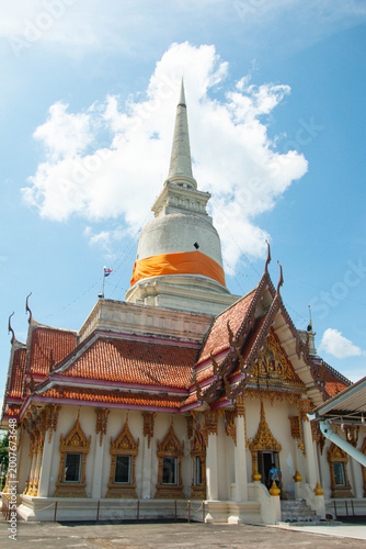 Traditional Thai temple with white stupa and orange cloth, viewed from low angle under bright blue sky and clouds, cultural landmark architecture.
