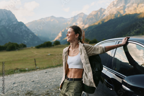 A woman beside a car in a mountainous landscape, wearing a white top and casual jacket, conveys outdoor freedom, travel lifestyle, and calm, confident mood.