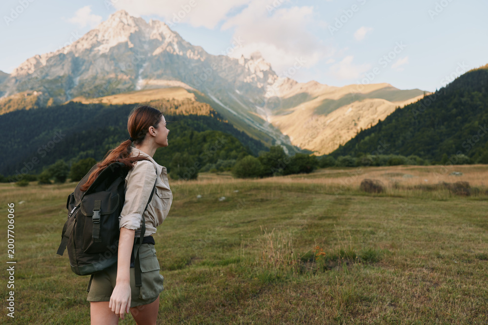 Naklejka premium A woman hiker with a backpack stands in an open meadow, facing distant mountains, capturing a moment of outdoor adventure and solo nature travel. In bright daytime scenery.