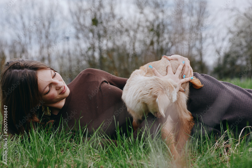Naklejka premium Woman lying on grass in a cosy sweater while a goat kid nudges her hand, capturing warmth, companionship, and rustic charm in an outdoor setting. Natural light creates a calm, lifestyle mood.