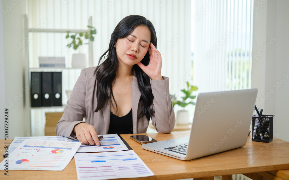 Fototapeta premium Stressed businesswoman holding her temples while working on a laptop, surrounded by financial documents in a modern office.