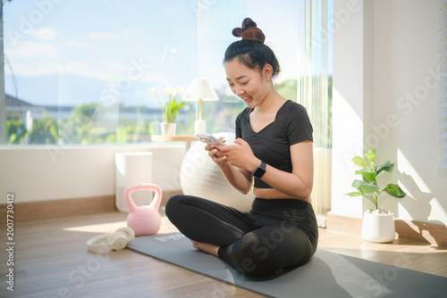 Young woman sitting on a yoga mat, checking her smartphone after a workout, surrounded by fitness equipment in a bright home. Free Download Young woman sitting on a yoga mat, checking her smartphone after a workout, surrounded by fitness equipment in a bright home. - GFXLoad