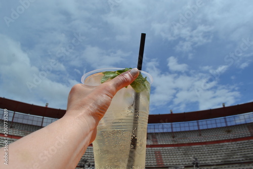 Obraz na plátně Hand holding rebujito cup against blue sky and bullring at feria de abril for fe