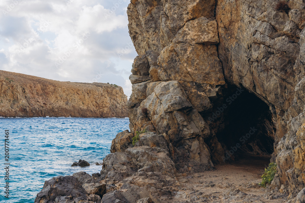 Fototapeta premium Rocky path leading into a natural sea cave on a cliff face, with turquoise Mediterranean Sea and distant headland