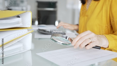 A professional female hand in a yellow blouse holding a magnifying glass and examining financial data on paper, checking details in accounting and finance. Audit and business taxes