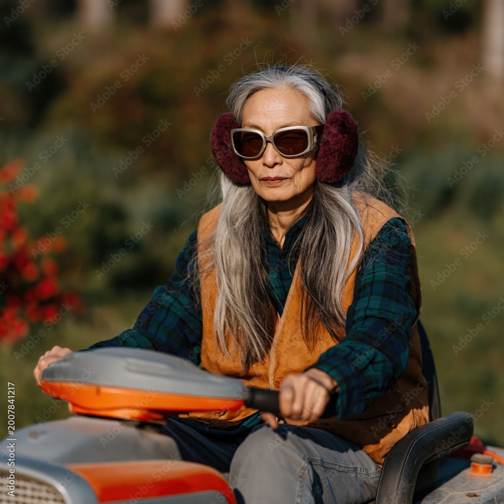 Obraz premium Older woman riding mower through landscaped yard wearing earmuffs