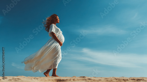 Pregnant woman walks on the beach during daytime with a clear blue sky and light wind moving her dress