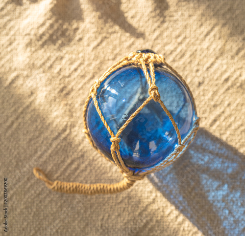 Tableau sur toile Blue glass fishing float wrapped in rope resting on sand with warm sunlight and coastal shadow patterns