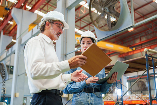 Two Caucasian men are meticulously examining metal sheets and cardboard box within warehouse environment, engaged in focused discussion with serious expression while inspecting production materials.