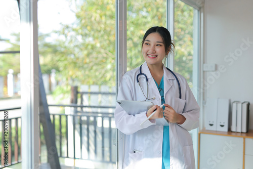 Obraz na plátně Young Asian female doctor in lab coat and stethoscope smiling, holding a digital