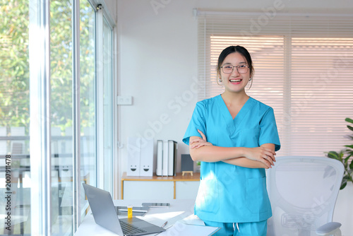 Smiling asian female doctor or nurse in a blue uniform standing in a clinic or hospital office, arms crossed