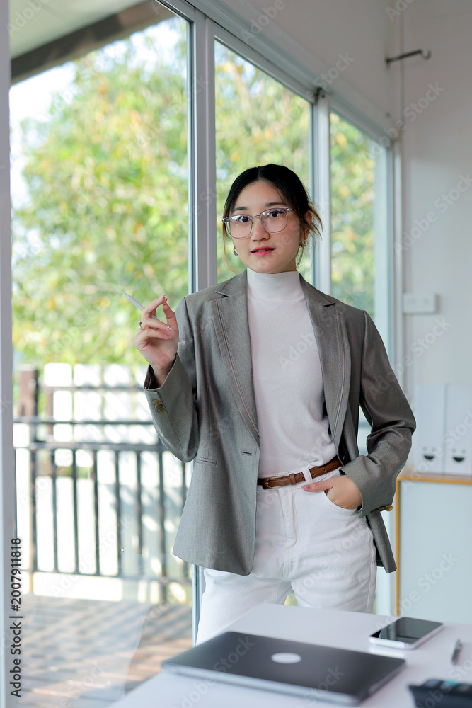 Fototapeta premium Young professional Asian businesswoman wearing glasses and blazer, standing by office window with hand in pocket, holding a pen