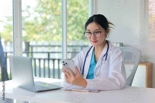 Young Asian female doctor in a lab coat and stethoscope holding a smartphone for a telemedicine video call in her office