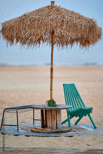 Foto Straw palapa umbrella with a wooden cable reel table and a teal wooden lounge chair on the sandy beach of Sopot, Pomeranian, Poland, Europe