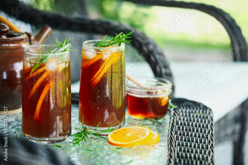 Cold ice herbal tea with orange slices and rosemary stem leaves in  tall glasses. Selective focus on second glass with blurred foreground and background..