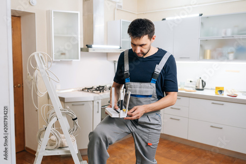 Focused technician in uniform installs network router for home internet. Man holding device in modern kitchen with stepladder and cables nearby. Professional service and connectivity concept.