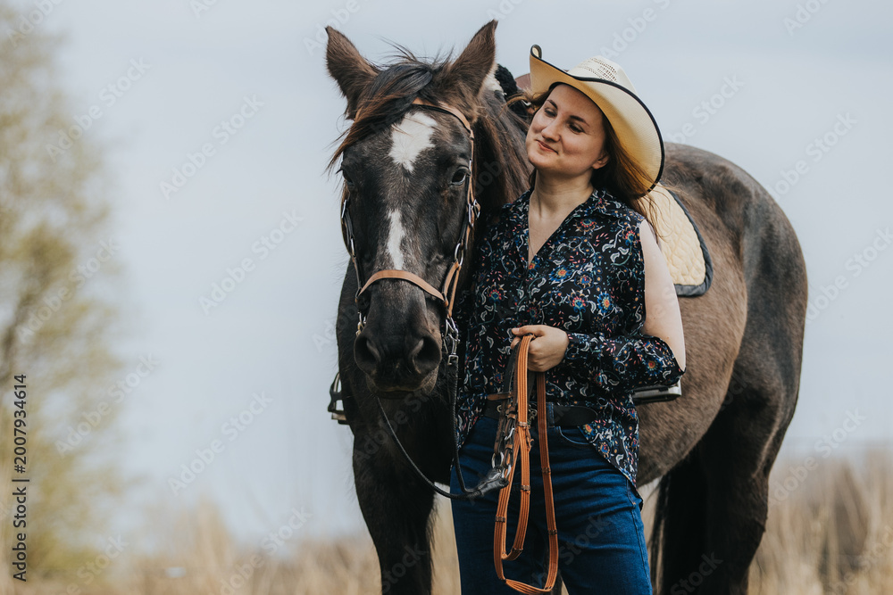 Obraz premium Young woman in floral shirt and cowboy hat stands beside brown horse holding reins in a grassy field with trees and cloudy sky in the background