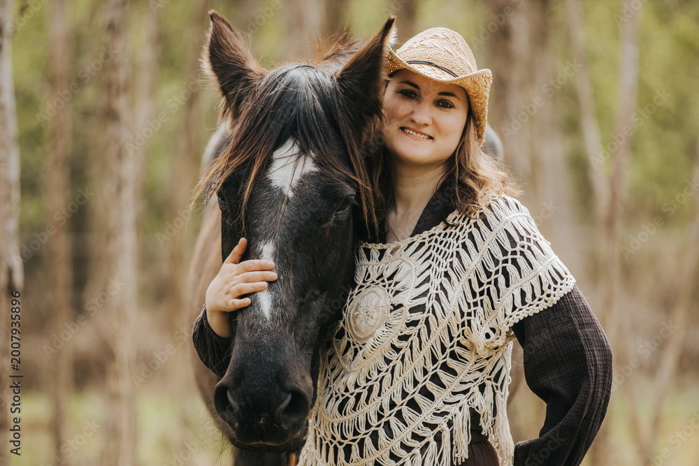 Obraz premium Young woman wearing a straw hat and crochet poncho stands beside a black horse in a forested area with trees and greenery in the background