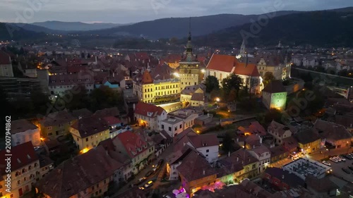 Nighttime drone footage of the illuminated medieval Clock Tower and old town of Sighisoara, Romania. Cinematic view of the historic citadel with city lights and evening atmosphere.