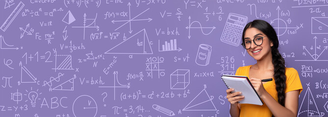 A student stands in a classroom setting with a purple collage of math and science drawings behind her. She is holding a notepad and writing while looking at the board. It is daytime.