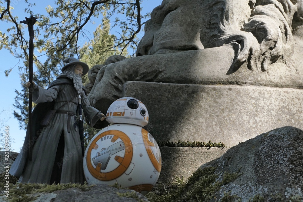 Fototapeta premium Action figure of fantasy wizard Gandalf known from novels Hobit and Lord Of The Rings, following spherical Star Wars astromech droid BB8 on mossy path next to huge stone lion statue.