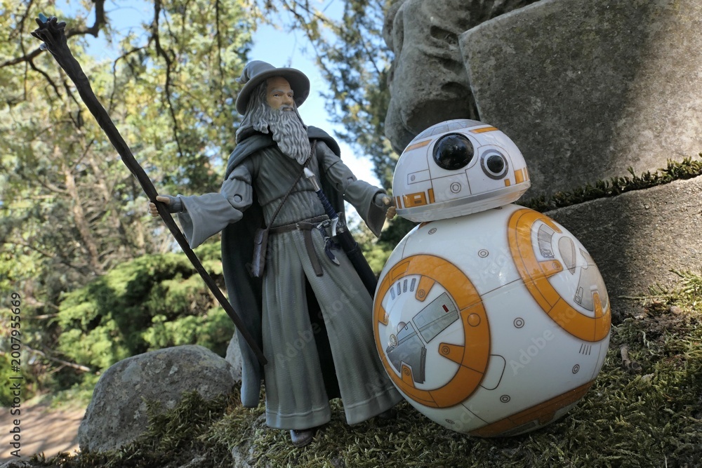 Fototapeta premium Action figure of fantasy wizard Gandalf known from novels Hobit and Lord Of The Rings, following spherical Star Wars astromech droid BB8 on mossy path next to huge stone lion statue. Park background. 
