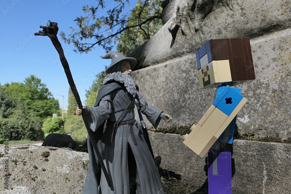 Naklejka premium Minecraft figure of main character Steve greets action figure of wizard Gandalf from novel Hobit and Lord Of The Rings by J.R.R.Tolkien under ancient stone pedestal of lion statue, spring sunshine. 