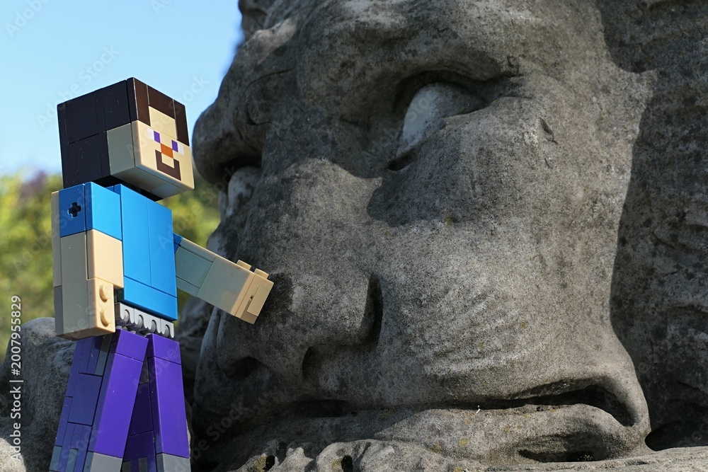 Naklejka premium Action figure of main Minecraft Character Steve standing next to a face of giant lion statue in park, spring blue skies in background. 
