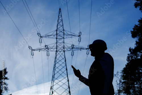Foto Silhouette of a male power engineer with a phone in his hand stands against the background of a power line and a blue sky