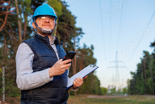 Energy industry expert performing a digital inspection of transmission towers. Male engineer recording maintenance data on a mobile app near a power grid pylon during site survey.