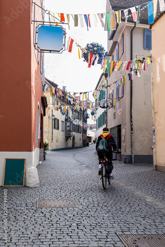Morning street scene in Konstanz, Germany with a cyclist riding through a narrow old town street decorated with colorful flags.