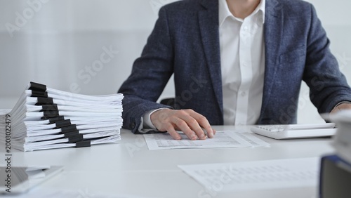 Businessman sorting through stacks of documents on a white desk beside a tall pile of bound papers, symbolizing meticulous financial record keeping and bureaucracy. Audit and taxes