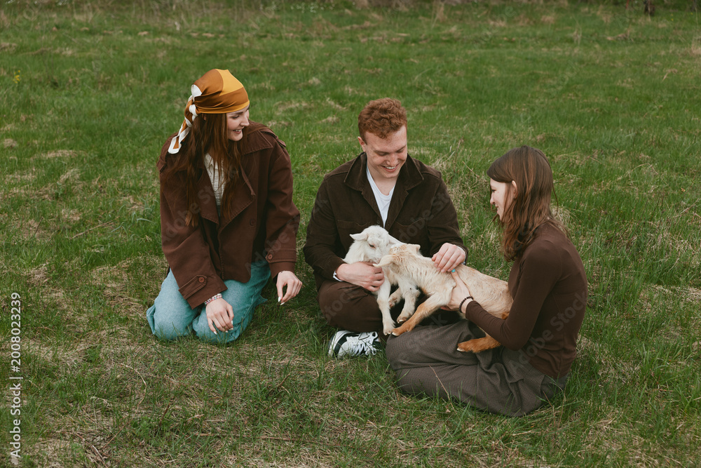 Naklejka premium Three friends outdoors share a gentle moment with a goat kid, sitting on grass, smiling, petting, sunlit scene, casual clothing, peaceful rural vibe, natural colors, candid lifestyle
