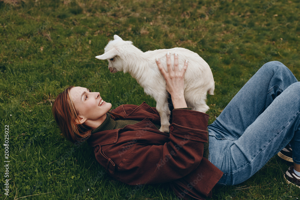 Naklejka premium Young woman lying on grass holds a playful white baby goat above her while smiling, enjoying a relaxed outdoor moment in casual clothing during a lifestyle photoshoot.