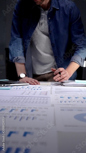 Entrepreneur standing at a desk in a dark office, studying financial reports and charts by lamp light while working on a laptop, focused on strategic planning and data analysis