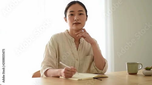 Young asian woman in cream linen shirt at light-wood desk, scribbling in notebook, lifts head with serene smile and reflective gaze. Sun-drenched modern apartment, soft bokeh. Dolly push-in from