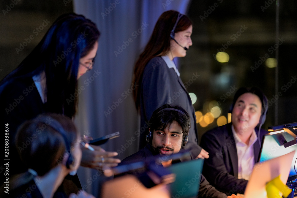 Fototapeta premium Multiethnic corporate colleagues working late. Professional female manager standing and assisting male operator wearing headset in a modern office with blurred foreground.