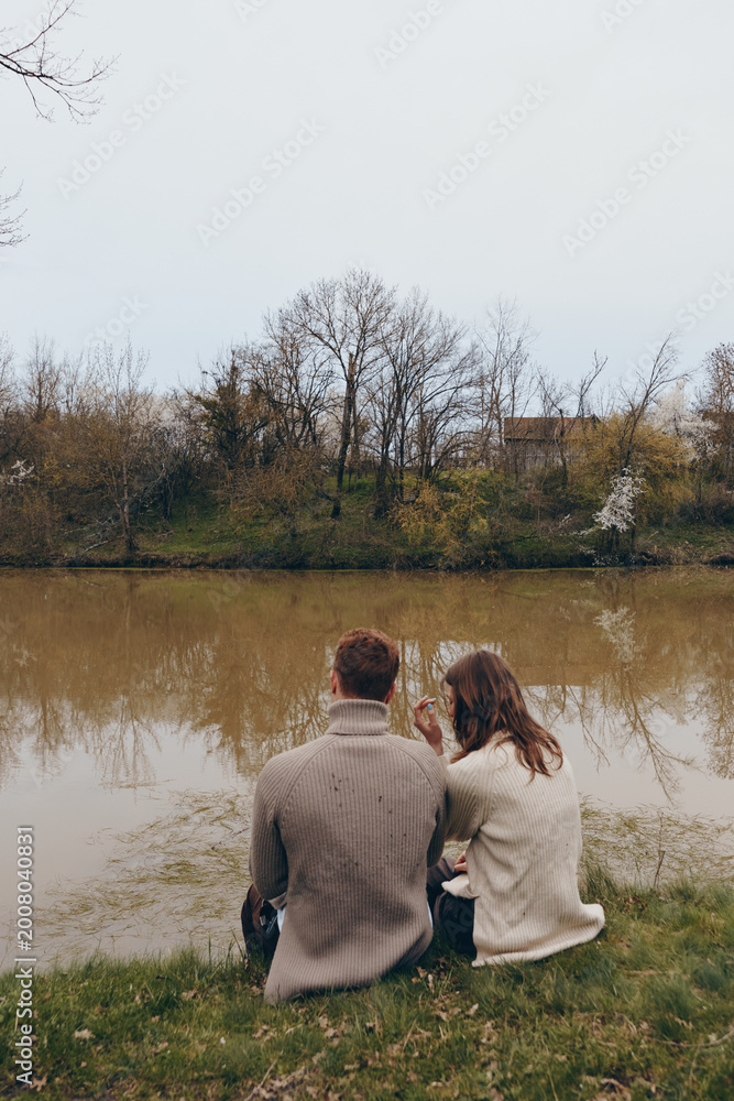Fototapeta premium Couple sitting by a calm lake in autumn, bundled in knit sweaters, sharing a quiet moment. Warm tones, soft light, and a gentle breeze create a peaceful mood about connection and nature.