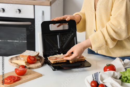 Photos Young woman using sandwich maker at table in kitchen