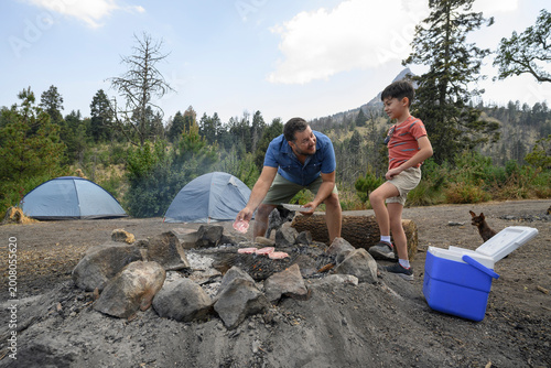 Father and son cooking meat on campfire grill while camping with dogs, family bonding in nature.