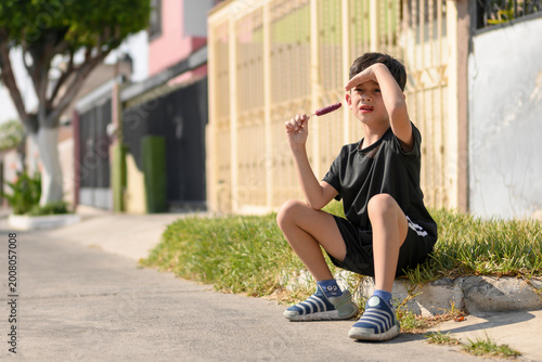 Little boy sitting on a sidewalk eating a popsicle and shading his eyes from the sun, summer in the city.