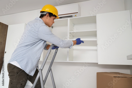 Fotografie Male worker assembling cupboard with shelves on stepladder in kitchen