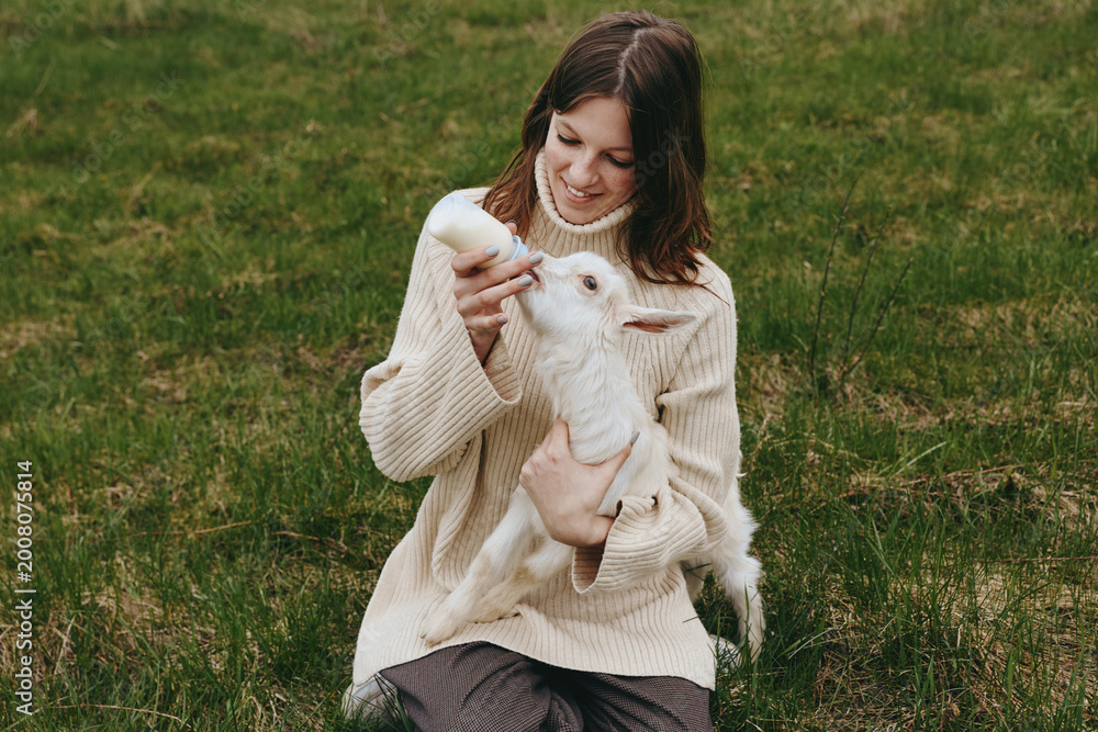 Naklejka premium A woman in a chunky beige sweater holds a white goat kid in a grassy field, sharing a tender moment of affection outdoors, showcasing rural lifestyle, animal companionship, and simple farm life.