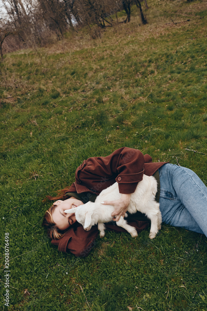 Naklejka premium Young woman lies on grass in casual jacket and jeans, cuddling a gentle goat kid, sharing a tender moment in nature, outdoor lifestyle scene with peaceful rural ambiance