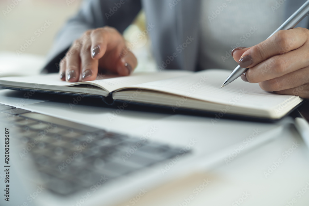 Fototapeta premium Business woman writing in notebook at office with laptop on desk, business planning, doing online research, close up