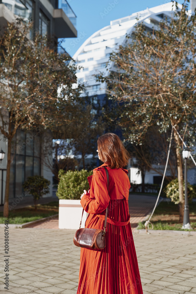 Fototapeta premium Young woman with red hair wears a long red dress and carries a brown leather bag while walking outdoors in a sunny urban park with autumn trees around her.