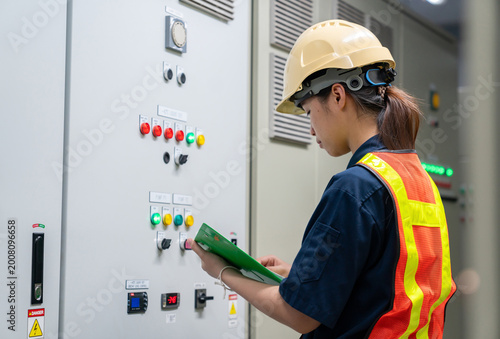 Professional female safety officer or electrical engineer in a hard hat and reflective vest inspecting a main power control panel at an industrial facility.