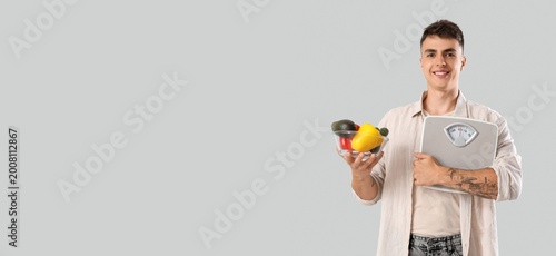 Young man holding weight scales and vegetables on grey background with space ...